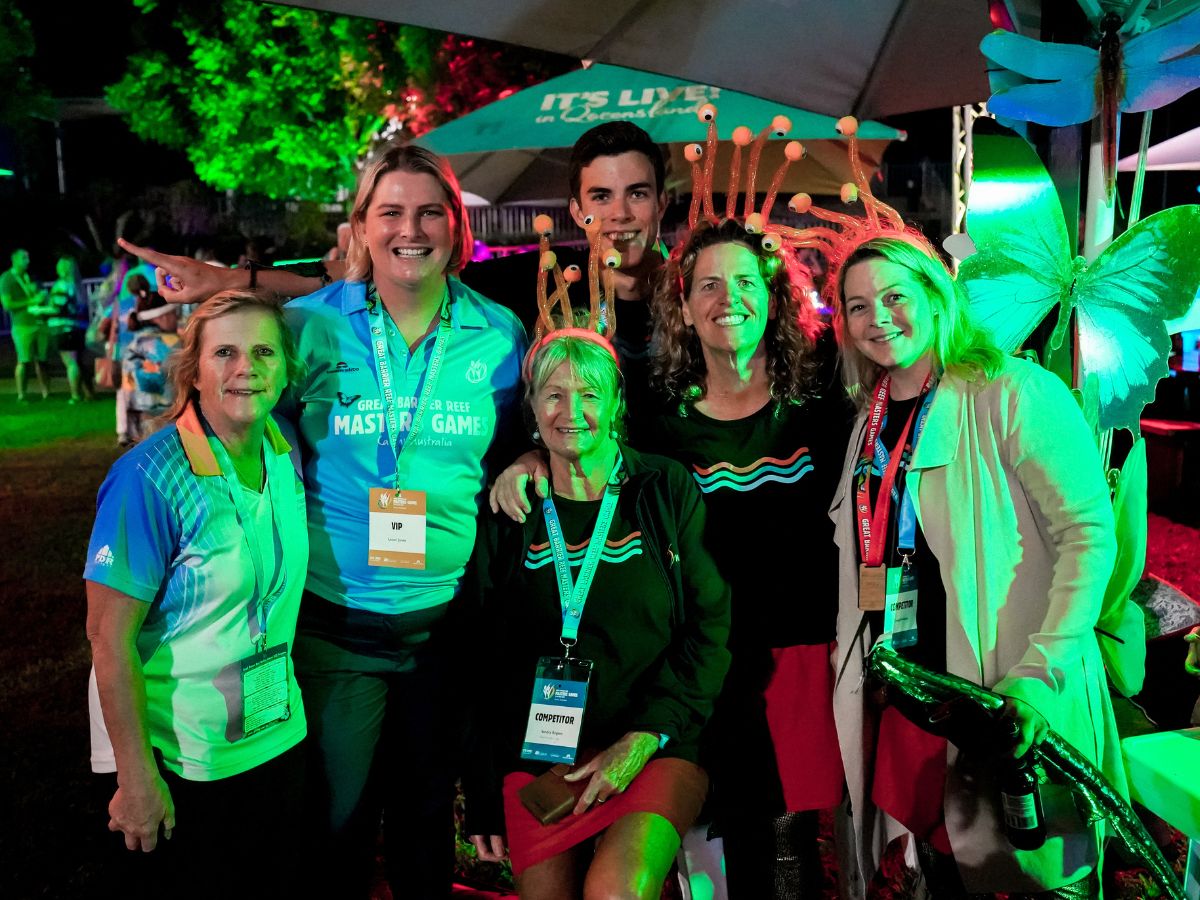 A group of masters competitors smiling and posing for a photo with vibrant, colorful lighting and decorations in the background