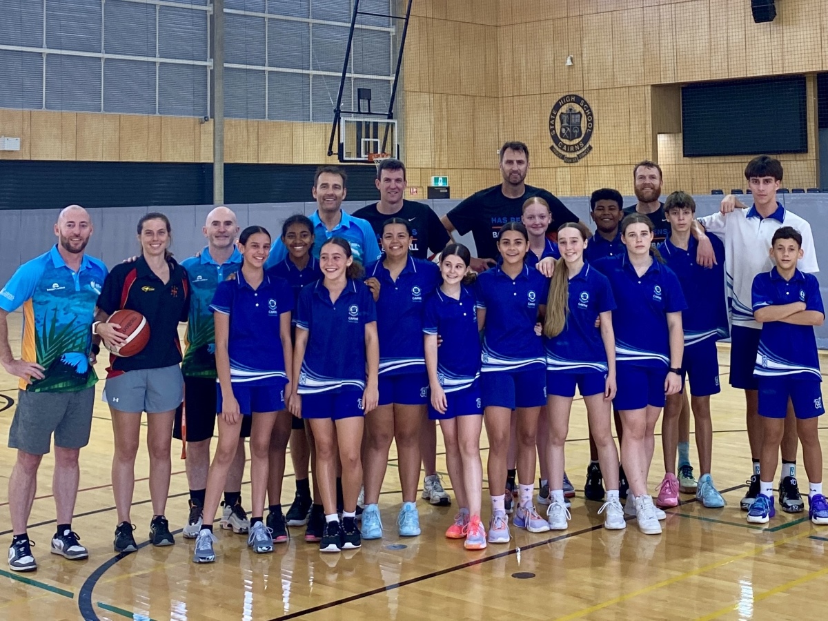 A group of students in sports wear are gathered in a gym for a basketball clinic